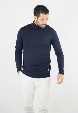 Pull col roulé bleu marine en tissu tricoté, avec des manches longues, des poignets côtelés et un petit logo sur l'ourlet. Porté avec un pantalon blanc.