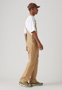 Salopette beige à jambes larges, avec bretelles réglables, poches avant et une légère texture. Associée à un t-shirt blanc et une casquette marron.