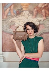 Žena v zelených šatech drží hnědou kabelku u obličeje, stojí před detailními zdobenými nástěnnými malbami v tlumených barvách.