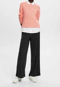 Korallinvärinen neulospusero valkoisen kauluspaidan päällä, yhdistettynä leveisiin mustiin housuihin ja valkoisiin lenkkareihin luoden kerroksellisen, rentoan tyylin.