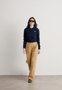 Námornícka polokošeľa s dlhým rukávom a golierom, béžové nohavice s vysokým pásom a čierne slnečné okuliare. Model stojí proti neutrálnemu pozadiu.