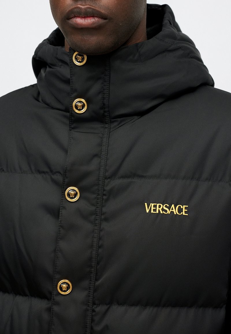 Sort polstret jakke med hætte, der har guldknapdetaljer og "VERSACE" broderet i guld på brystet. Glat, mat tekstur.