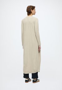 Beżowy długi kardigan o luźnym kroju, z prążkowanymi mankietami i bocznymi rozcięciami, wykonany z miękkiego materiału. Noszony na ciemnych spodniach i butach w paski.