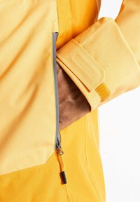 Gelbe Jacke mit glatter Textur, ausgestattet mit einer Reißverschlusstasche und einem verstellbaren Bündchen mit Klettverschluss. Detaillierte Nähte sind sichtbar.