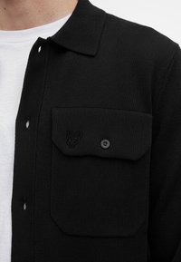 Veste noire en maille avec une poche poitrine boutonnée et un petit détail brodé de tête d’animal sur un t-shirt blanc à col rond.