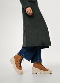 Bruine suède enkellaarzen met zwarte veters, een chunky beige zool en zichtbare stiksels, gecombineerd met blauwe wijde spijker Jeans en een donkere jas.