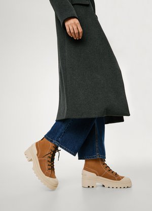 Brown suede ankle boots with black laces, a chunky beige sole, and visible stitching, paired with blue flared denim jeans and a dark coat.