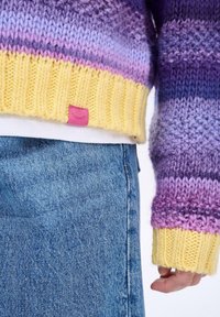Hrubý pletený sveter s gradientom fialovej, ružovej a žltej farby. Žlté žebrované manžety a lem; má malú ružovú etiketu. Kombinovaný s modrým denimom.
