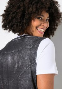 Chaleco de mezclilla negro con un acabado texturizado, combinado con una camiseta blanca de manga corta. Detalles de costura notables y una ligera curva en el hombro.
