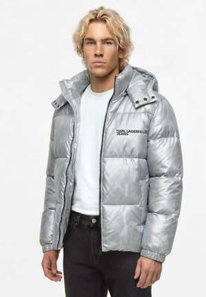 Ung mand med blondt hår iført en sølvfarvet dunjakke med hætte og "Karl Lagerfeld Jeans"-logo, hvid skjorte og sorte jeans.