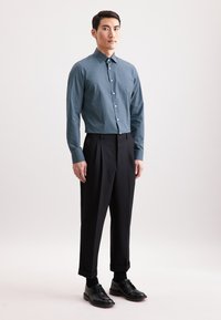 Camicia a maniche lunghe blu a fantasia con colletto, abbinata a pantaloni neri plissettati e scarpe di pelle nere, in piedi contro uno sfondo neutro.