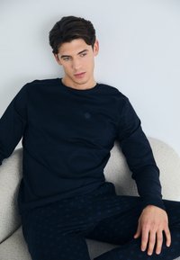 Marine lange mouwen shirt met een subtiele logo en bijpassende gestreepte broek, zittend op een gestructureerde lichtgekleurde stoel.