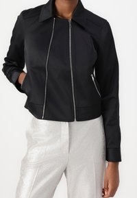 Veste noire courte avec une fermeture éclair à l'avant, deux poches latérales et une texture lisse. Elle présente un col pointu et des épaules légèrement structurées.