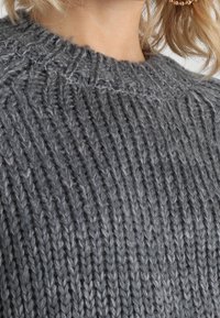 Pull gris tricoté avec une texture côtelée, présentant un col rond et un fil épais. Gros plan montrant des motifs de points distincts.