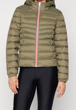 Personne portant une veste matelassée à capuche vert olive avec une fermeture éclair avant doublée de rose et des leggings noirs, debout devant un fond uni.