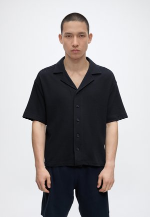 Tânăr cu păr scurt purtând o cămașă texturată neagră cu mâneci scurte și pantaloni scurți asortați, stând în fața unui fundal alb simplu.