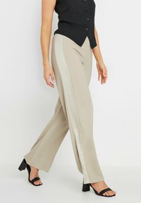 Pantalons larges beiges avec une rayure latérale de beige clair contrastante, fabriqués en tissu lisse, associés à des sandales à talons hauts noires.