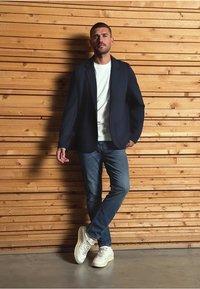 Marineblazer über einem weißen Hemd, kombiniert mit blauen Jeans und weißen Turnschuhen. Das Model posiert vor einer Holzwand mit horizontalen Latten.