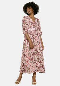 Maxi-jurk in een roze bloemenprint met korte pofmouwen, V-hals, een getailleerde taille en vloeiende stof. Draagt lichte sandalen.