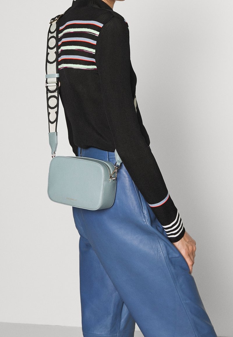 Jasnoniebieska skórzana torba na ramię o prostokątnym kształcie, z wzorzystym paskiem i subtelnym detalem logo. Noszona z czarnym swetrem w paski.