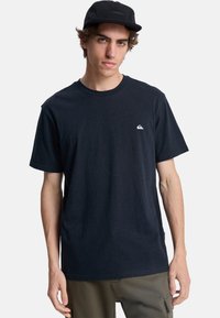 Camiseta negra de algodón, con cuello redondo, mangas cortas y un pequeño logo blanco en el lado izquierdo del pecho. Textura moteada sutil.