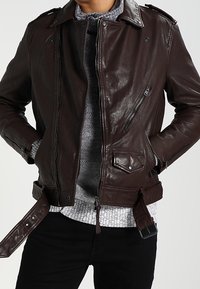 Bruine leren jas met een slim fit ontwerp, ritszakken en schouderepauletten. Sluit met een rits en heeft een riem aan de onderkant.