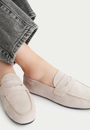 Mocasines de ante beige claro usados con pantalones vaqueros grises arremangados sobre un fondo neutro, destacando un estilo de calzado casual.