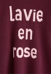 Burgundy stickad plagg med texten "Lavie en rose" i mjuk rosa. Texten är i en blockbokstavsfont, vilket skapar en kontrasterande visuell effekt på tyget.