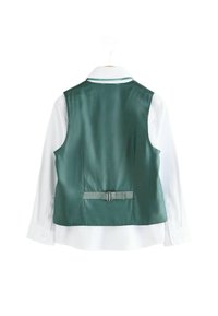 Grünes Satin-Weste mit einem taillierten Rücken, silberner Schnalle und einem weißen Langarmhemd, das am Kragen befestigt ist, hängt an einem weißen Haken.