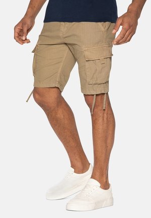 Beige Cargo-Shorts mit mehreren Taschen, aus strukturiertem Stoff, mit Kordelzug am Saum und lockerer Passform. Kombiniert mit weißen Turnschuhen.