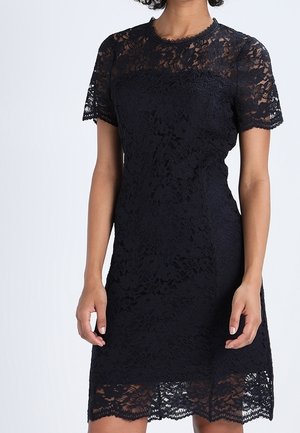 Femme portant une robe en dentelle noire longueur genoux avec des manches courtes et un ourlet festonné, debout devant un fond uni.