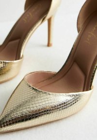Scarpe col tacco alto dorate con una finitura a serpente testurizzato, punta affusolata e design aperto sul retro. L'interno presenta una morbida fodera rosa.