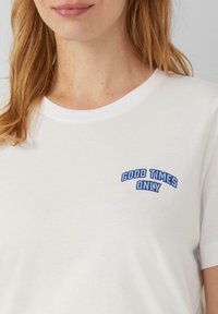 T-shirt en coton blanc à manches courtes et col rond côtelé. Présente un texte brodé bleu indiquant "QUE DES BONNES CHOSES" sur le devant.