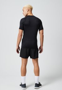 Zwarte korte mouwen sportshirt in combinatie met zwarte shorts. Het materiaal lijkt glad en aansluitend. Het model draagt witte sportkousen en sneakers.