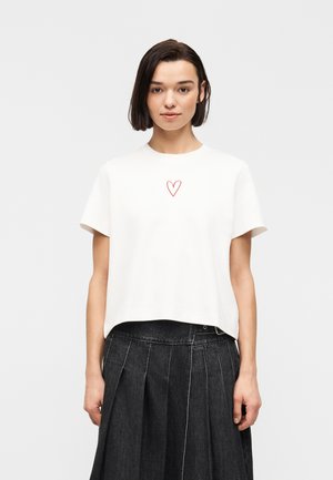 Γυναίκα με ίσια σκούρα μαλλιά που φοράει λευκό κοντομάνικο τοπ με μικρή κόκκινη καρδιά και μαύρη πλισέ φούστα.