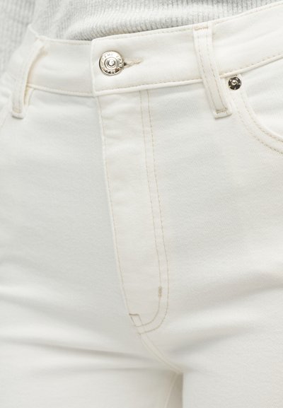 Κοντινή λήψη του μπροστινού μέρους λευκών τζιν από τζιν με ασημένιο κουμπί, θηλιές για ζώνη και ανοιχτό μπεζ ραφές, φορεμένα με λευκό ριμπ τοπ.