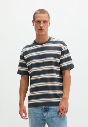 Man met kort haar, gekleed in een zwart-wit-grijs horizontaal gestreept T-shirt en blauwe spijkerbroek, staand tegen een effen witte achtergrond.