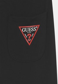 Svart stofflomme med en rød trekantet lapp som deler teksten "GUESS" sammen med et spørsmålstegnlogo, på en teksturert overflate.