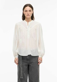 Valkoinen, pitkähihainen pusero, jossa on nappikiinnitteinen etulista, olkapäillä laskostetut yksityiskohdat ja rento istuvuus. Yhdistetty tummanharmaisiin, leveälahkeisiin housuihin.