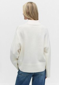 Vit stickad tröja med hög krage, nedhasade axlar och ribbad fåll. Parat med blå denimjeans, sedd bakifrån.