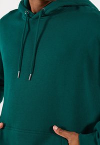 Vihreä huppari, valmistettu pehmeästä puuvillasta. Siinä on kengurutasku, kiristysnauhalla varustettu huppu metallisilla päillä sekä resorit hihansuissa ja helmassa.