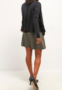 Žena oblečená v černém topu s dlouhým rukávem, olivově zelené sukni délky ke kolenům a černých vysokých podpatcích stojící před bílým pozadím.