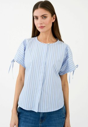 Žena nosi svetlomodrú blúzku s krátkym rukávom, vertikálne pruhovanú, so zaväzovaním na rukávoch, v kombinácii s modrými džínsami, stojaca na bielom pozadí.