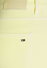 Tejido de denim amarillo claro con costuras blancas y una etiqueta de cuero de Tommy Hilfiger. Presenta una pequeña etiqueta de logo multicolor en el bolsillo.