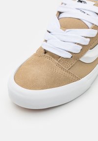 Tan suede sneaker with white laces, textured rubber sole, and a smooth toe cap. Features a contrasting white side stripe.