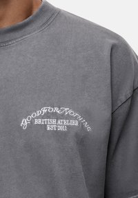 Szara bawełniana koszulka z okrągłym dekoltem, luźnym krojem oraz haftowanym logo w kolorze białym: "GOOD FOR NOTHING BRITISH ATELIER EST 2011."