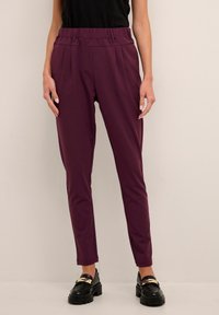Pantalon sur mesure bordeaux avec une texture lisse, taille élastique et design fuselé, associé à des chaussures noires ornées d'accents dorés.