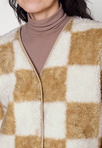 Beige och krämfärgad rutig fleeceväst med en mjuk, texturerad yta, som har en kontrasterande beige kant och en enkel, öppen framsidesdesign.