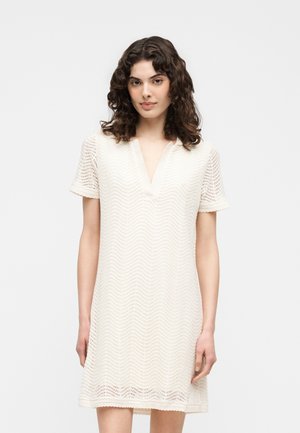 Femme aux cheveux bouclés foncés portant une robe blanche en dentelle à manches courtes avec un décolleté en V, debout sur un fond clair uni.