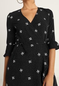 Schwarzes Kleid mit weißem Blumenmuster und Polka-Dots. Verfügt über einen V-Ausschnitt, Dreiviertelärmel und eine gebundene Taillenpartie.